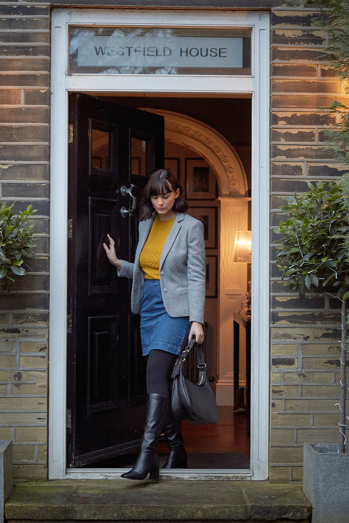 Woman exiting a building named 'Westfield House' wearing a gray blazer, yellow top, blue skirt, black opaque tights  and black boots.