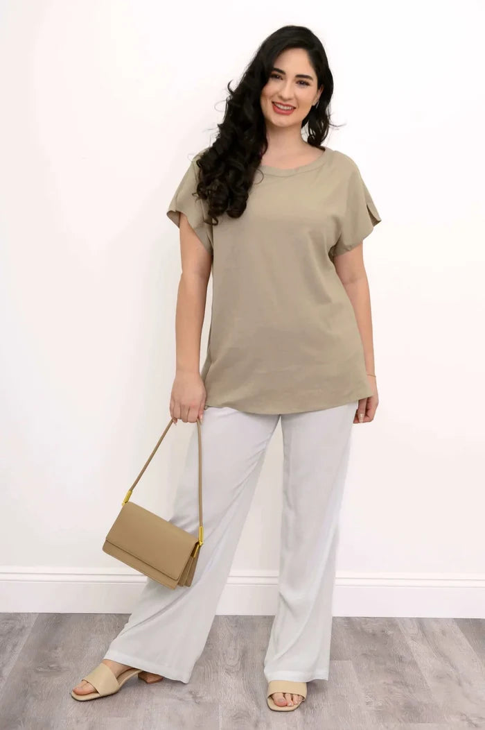 Woman wearing a beige top and white pants holding a beige handbag against a white background