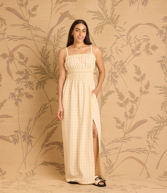 A woman modeling a light beige and white gingham maxi dress with spaghetti straps, a smocked bodice, and a high side slit, standing against a beige botanical patterned backdrop.