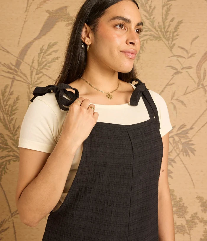 Woman wearing a black pinafore over a white shirt against a beige floral-patterned background