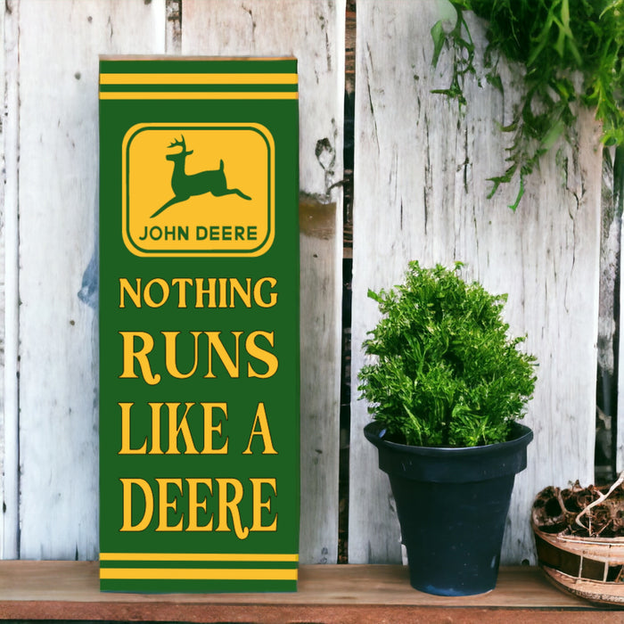 A vertical, retro style John Deere sign featuring the brand logo with the text 'NOTHING RUNS LIKE A DEERE' in bold lettering, placed against a wooden backdrop with green plants to the side.