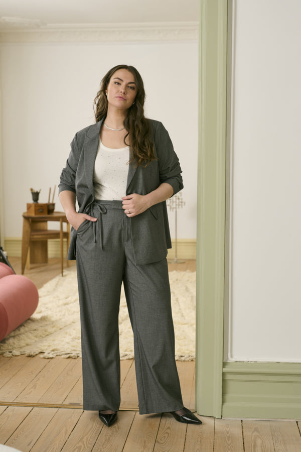 Woman in a gray outfit standing in a room with wooden flooring and a white wall.